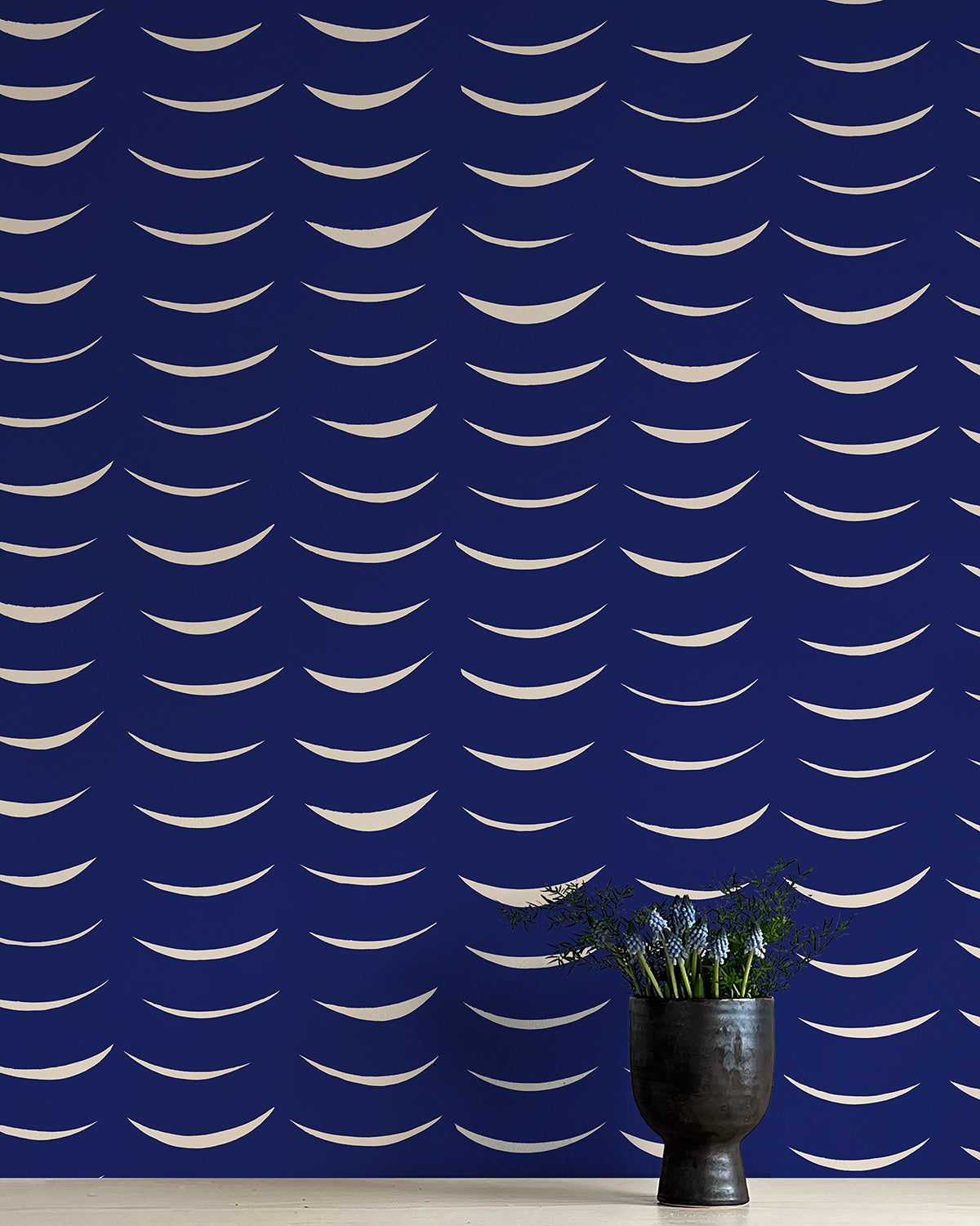 A vase of flowers stands in front of a wall papered in an abstract crescent print in cream on a navy field.