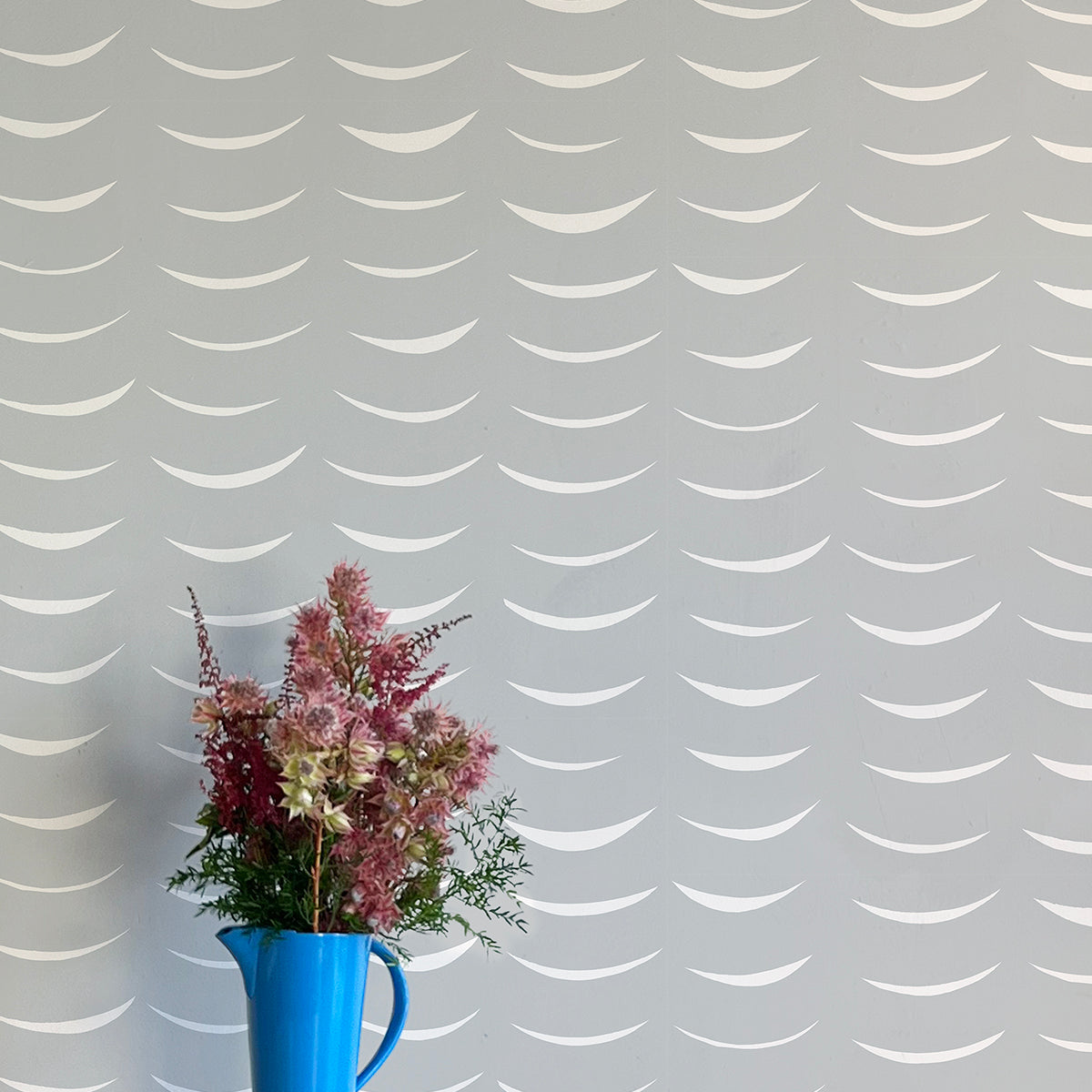 A vase of flowers stands in front of a wall papered in an abstract crescent print in cream on a gray field.