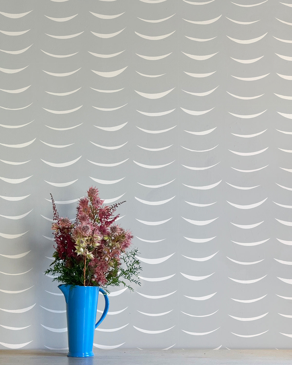 A vase of flowers stands in front of a wall papered in an abstract crescent print in cream on a gray field.