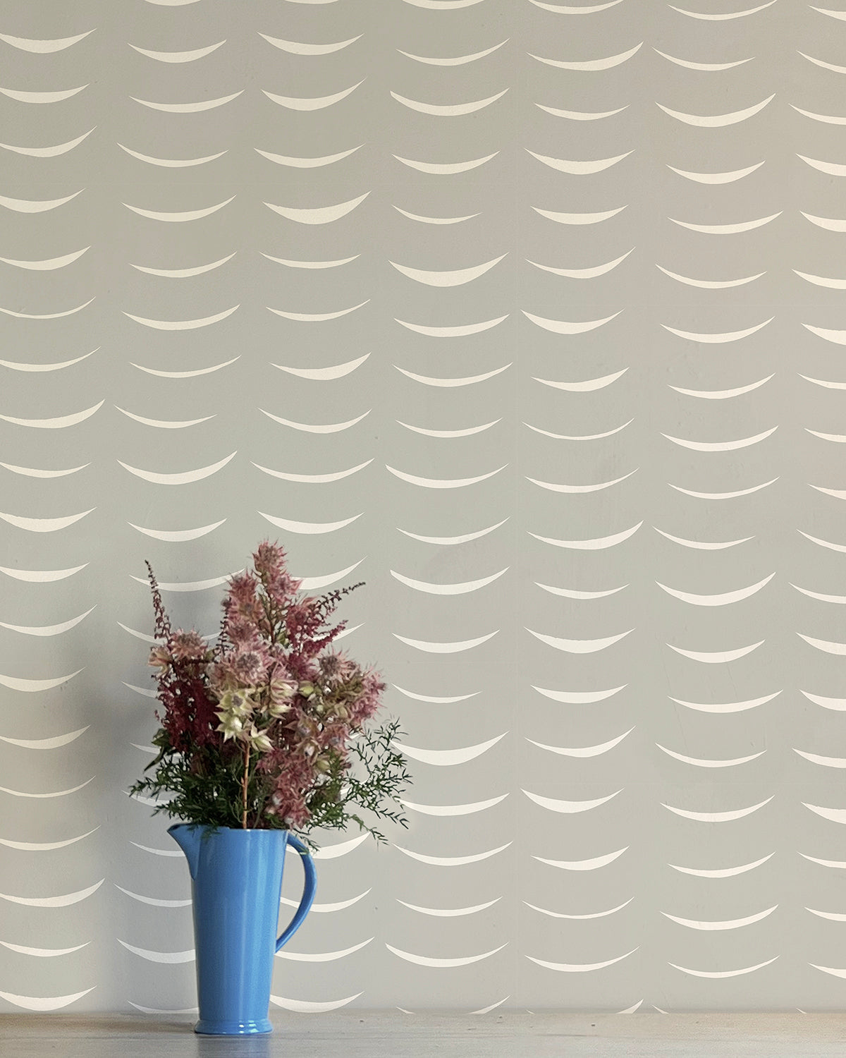 A vase of flowers stands in front of a wall papered in an abstract crescent print in cream on a gray field.