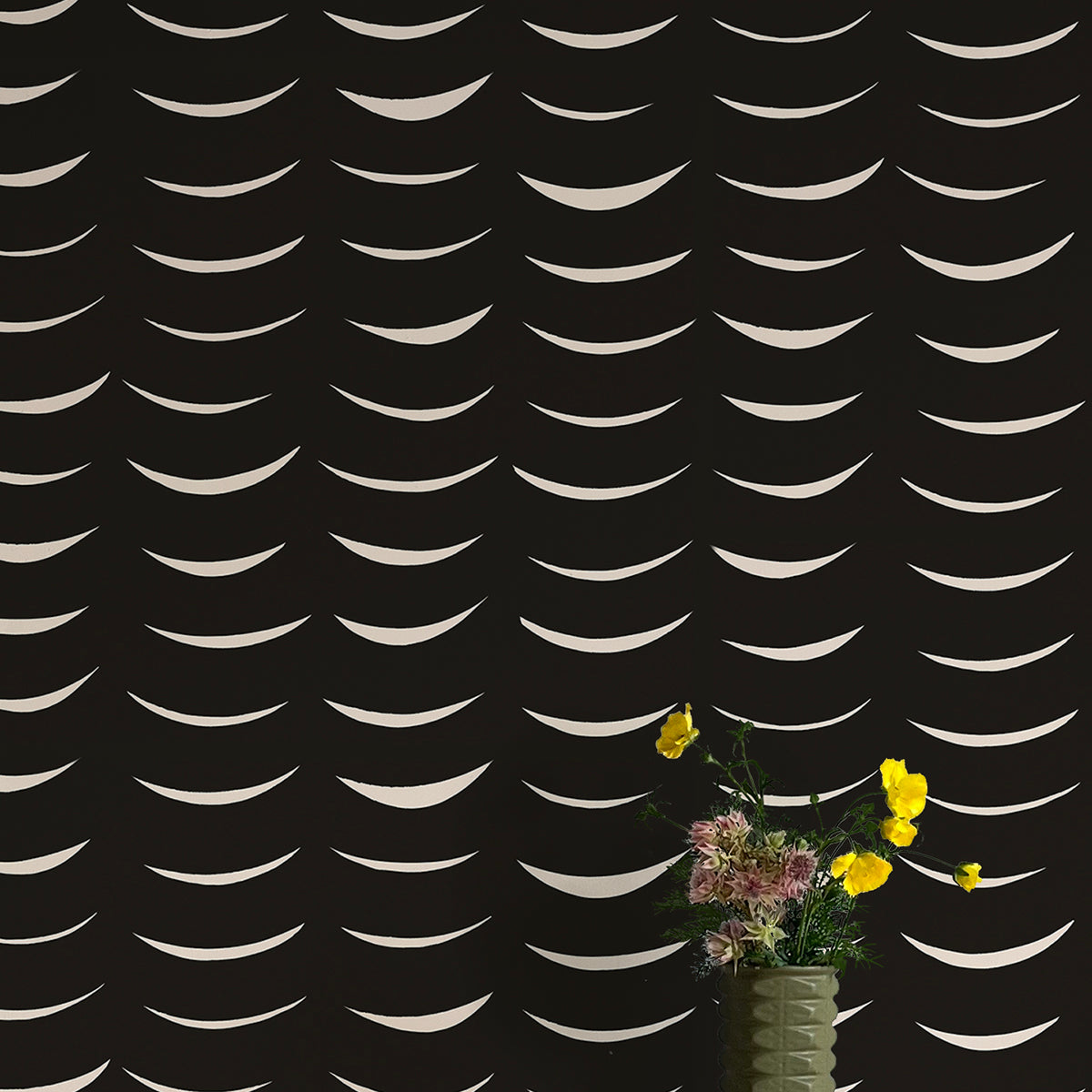 A vase of flowers stands in front of a wall papered in an abstract crescent print in cream on a black field.