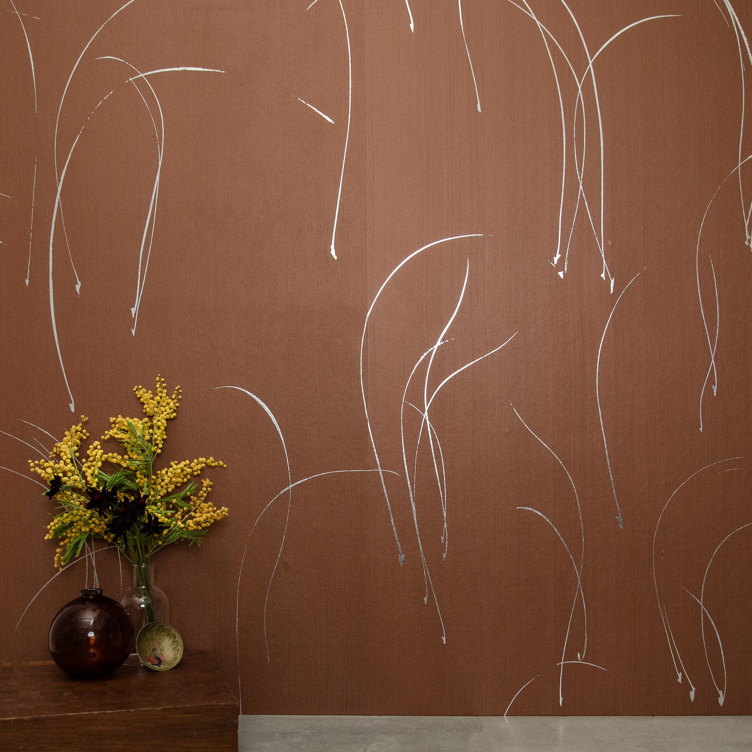 A vase of flowers stands in front of a wall papered in an elongated paint splatter pattern in silver on a brown field.
