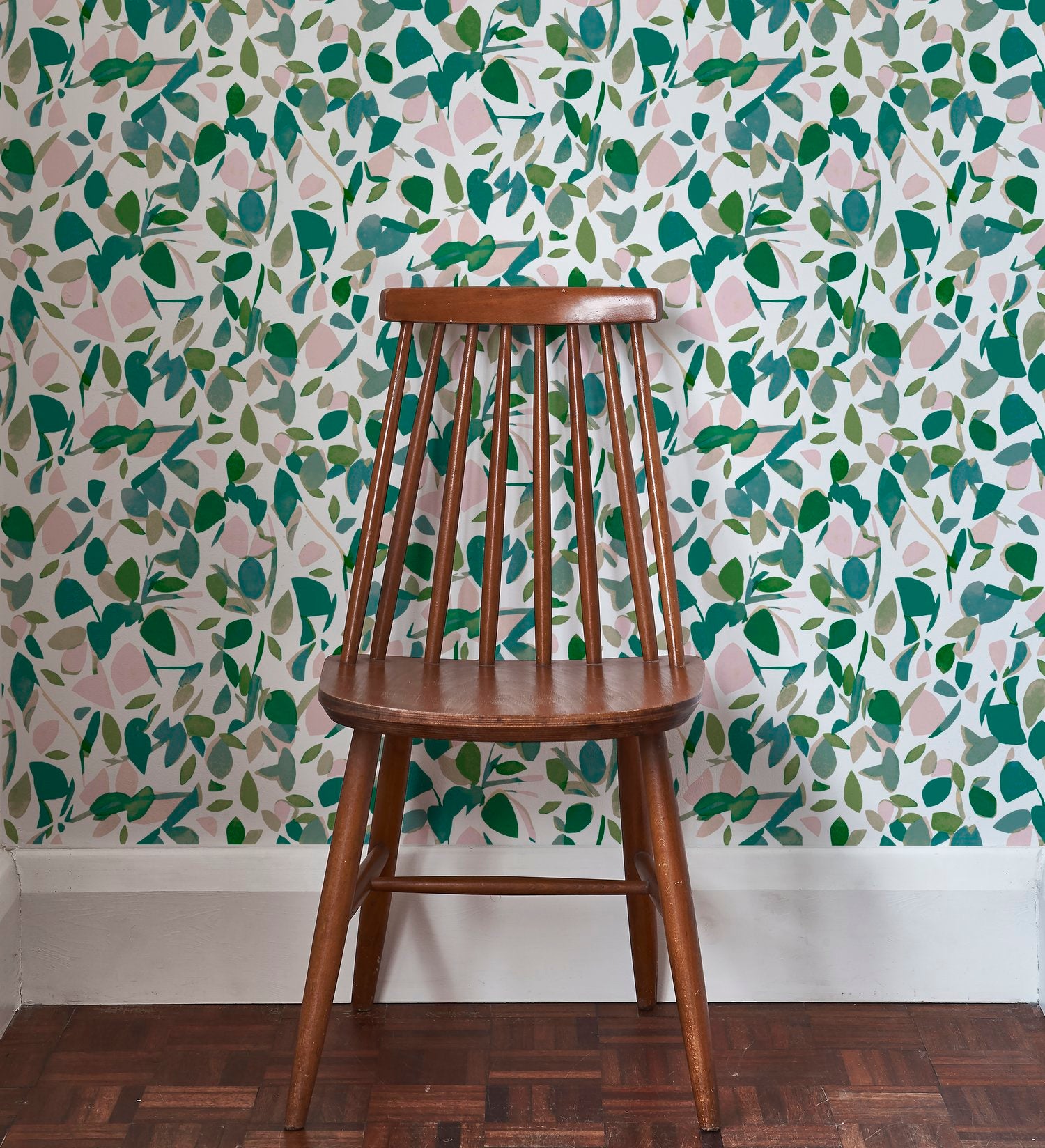 A wooden chair stands in front of a wall papered in a minimal leaf print in shades of green, pink and white.
