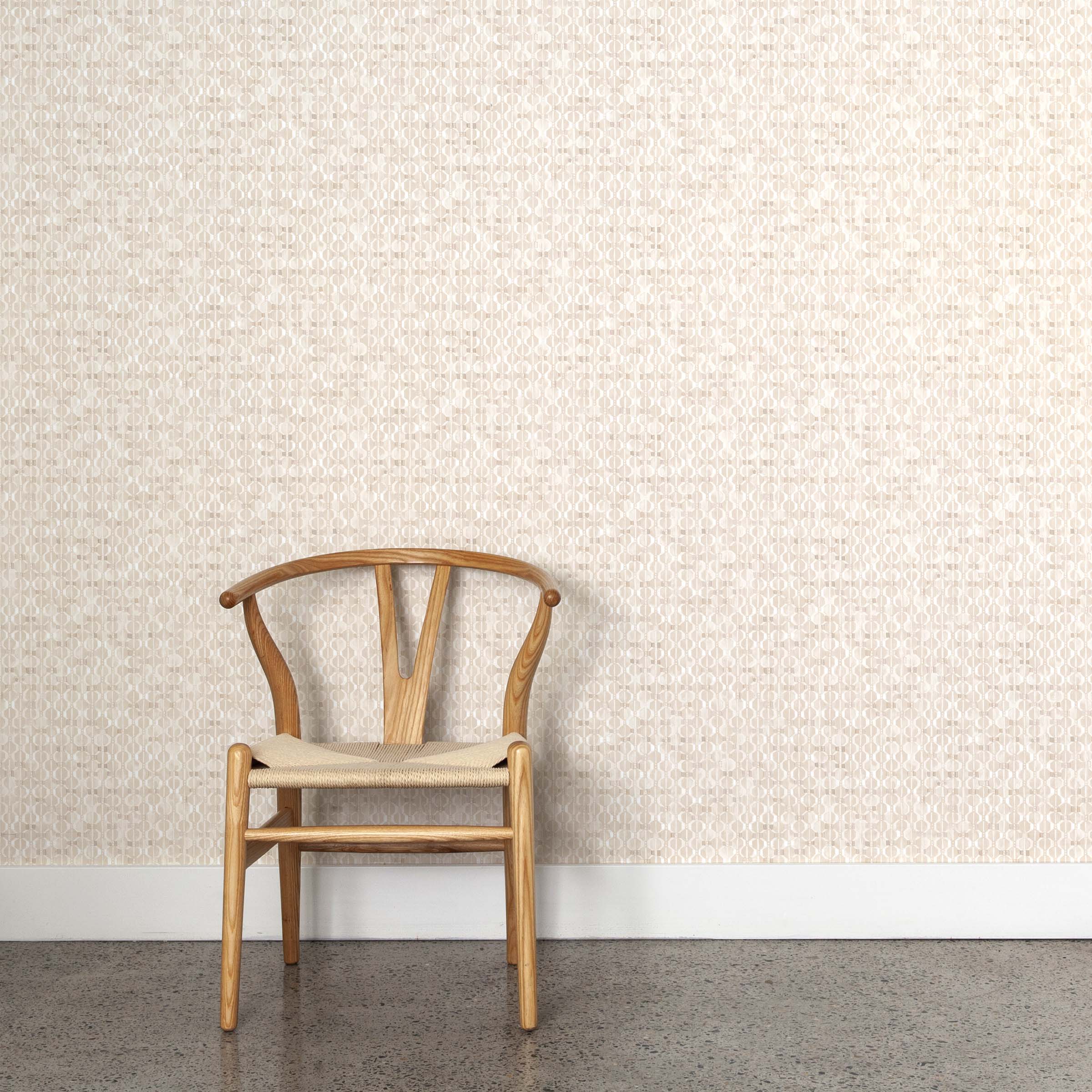 A wooden chair stands in front of a wall papered in a textural geometric print in shades of cream on a white field.