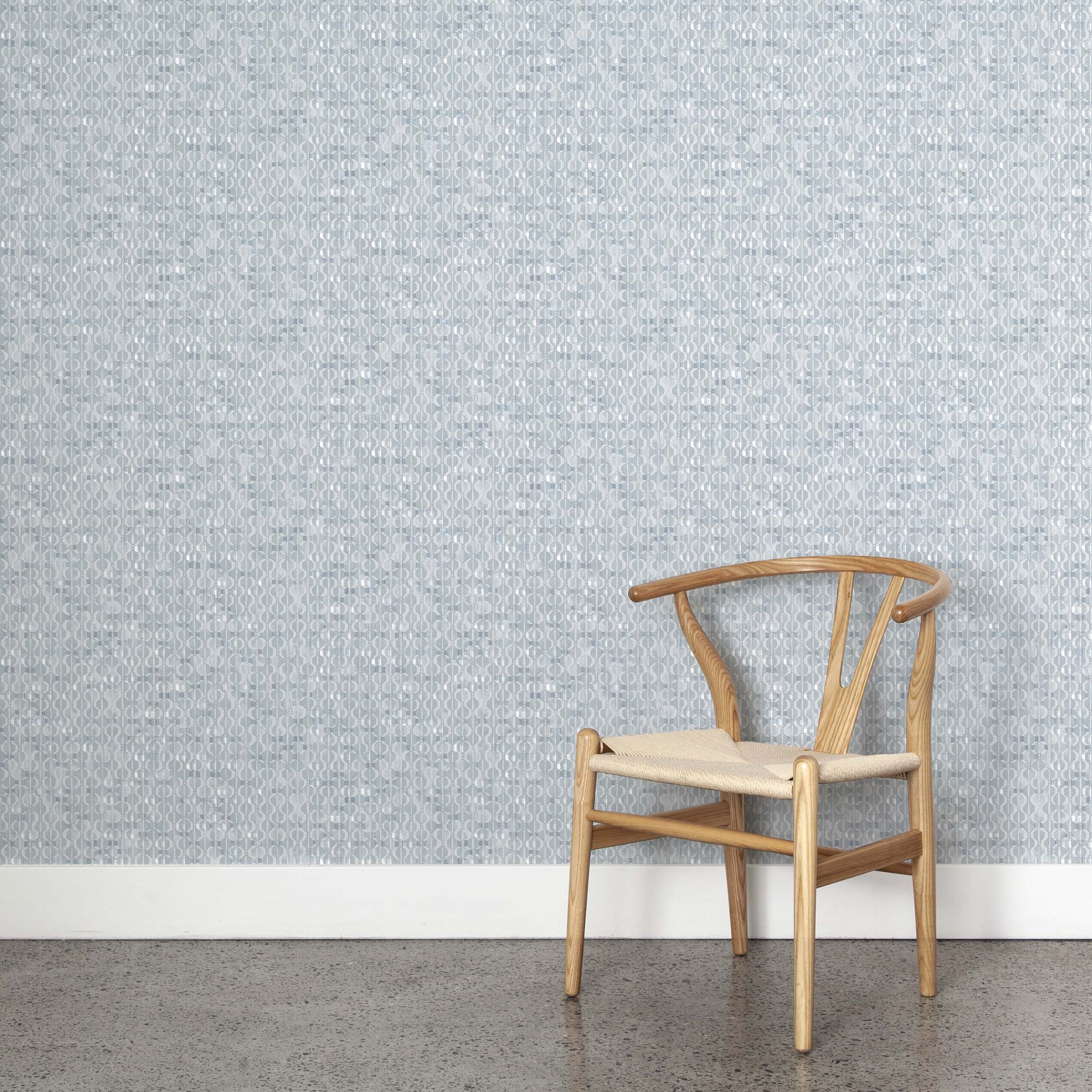 A wooden chair stands in front of a wall papered in a textural geometric print in shades of blue on a white field.