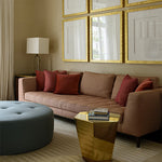 Living room scene with brown couch, gold side table, blue circular ottoman against neutral walls with gold framed art. 