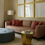 Living room scene with brown couch, gold side table, blue circular ottoman against neutral walls with gold framed art. 