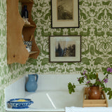 Bathtub with green patterned wallpaper, wooden shelf, and various items on the ledge.