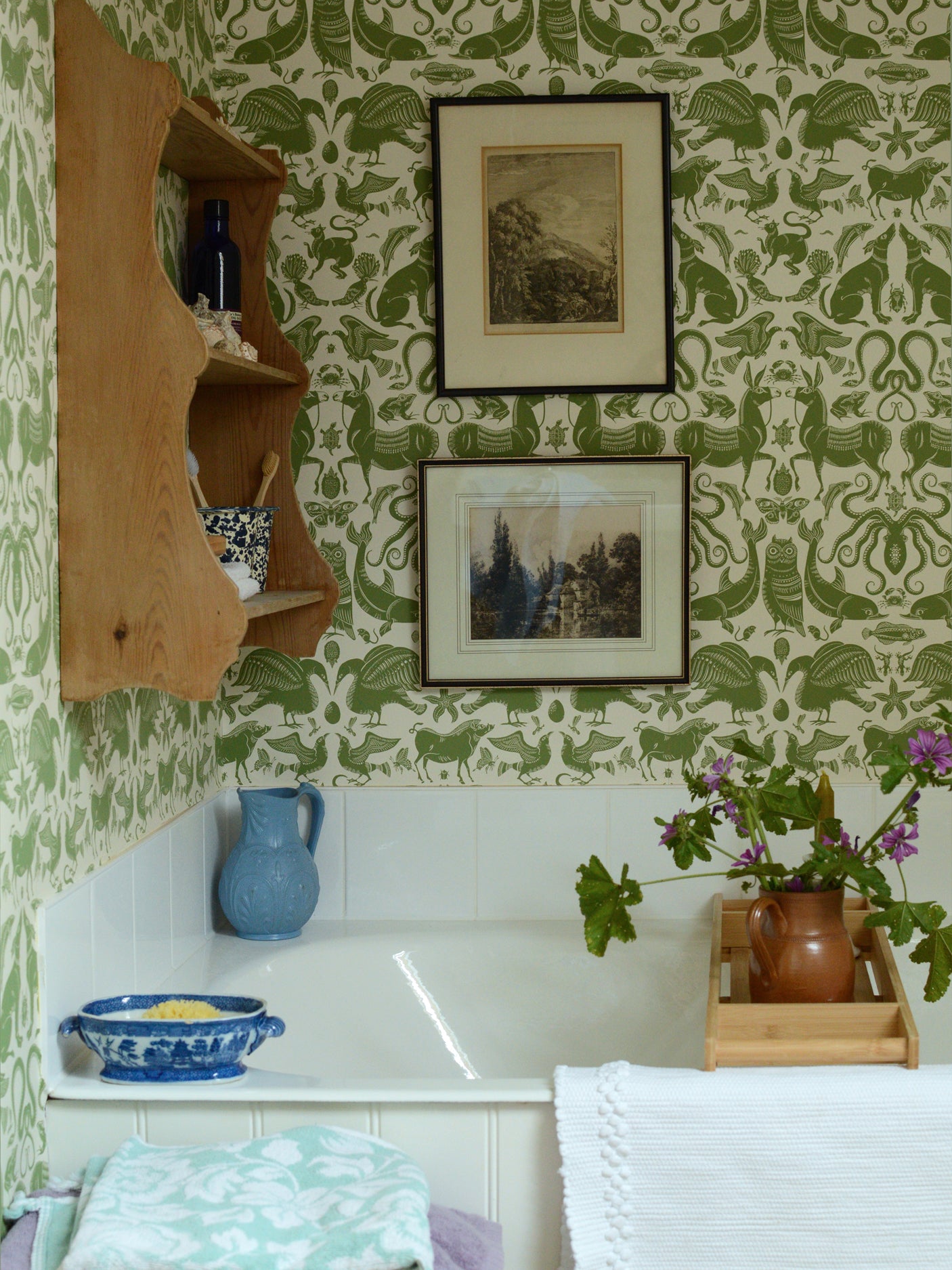 Bathtub with green patterned wallpaper, wooden shelf, and various items on the ledge.