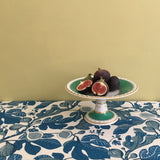 Decorative table setting with a cake stand holding figs on a tablecloth with fig design.