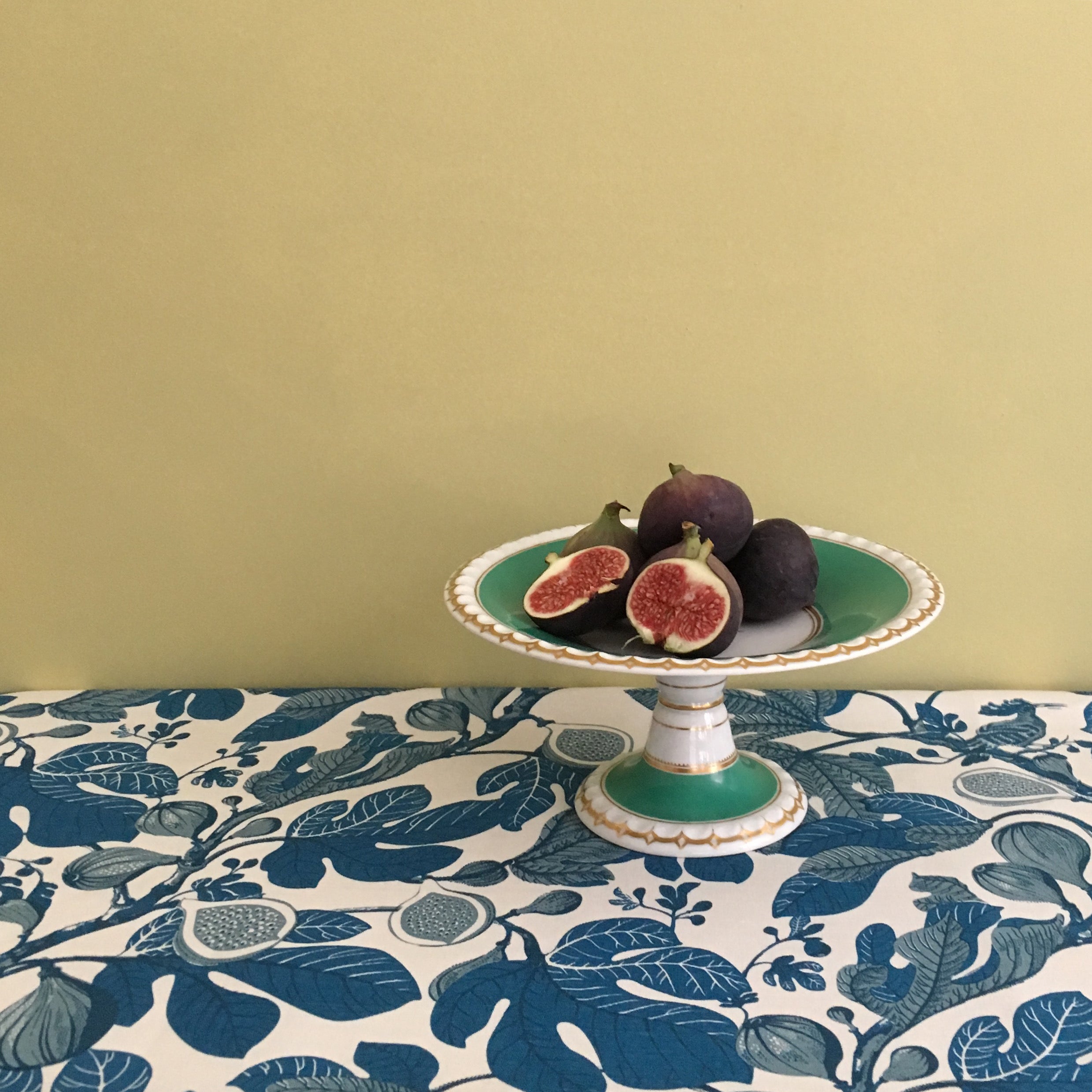 Decorative table setting with a cake stand holding figs on a tablecloth with fig design.