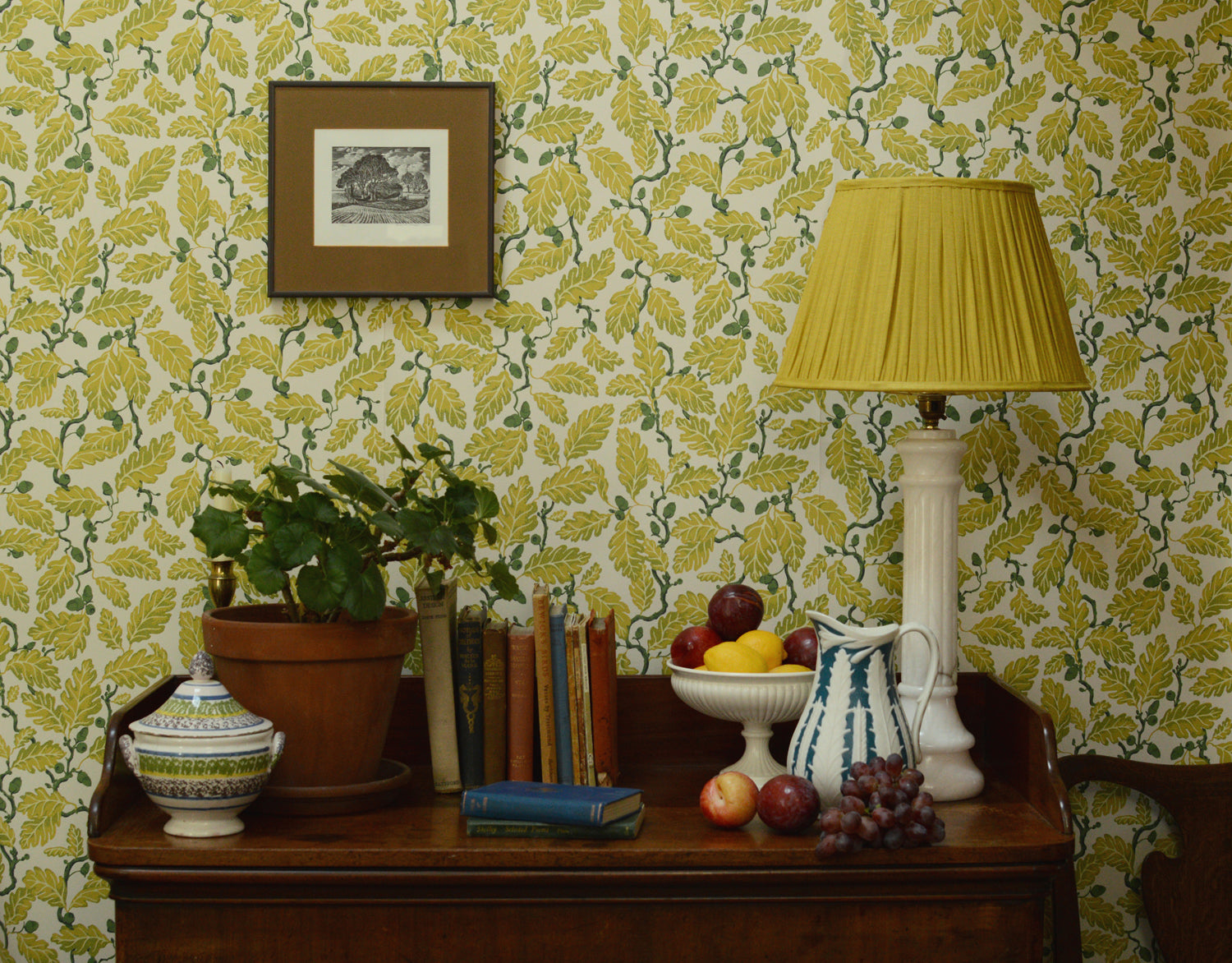 Room interior with a wooden sideboard against a patterned wall, featuring a lamp, books, fruit, and a plant.