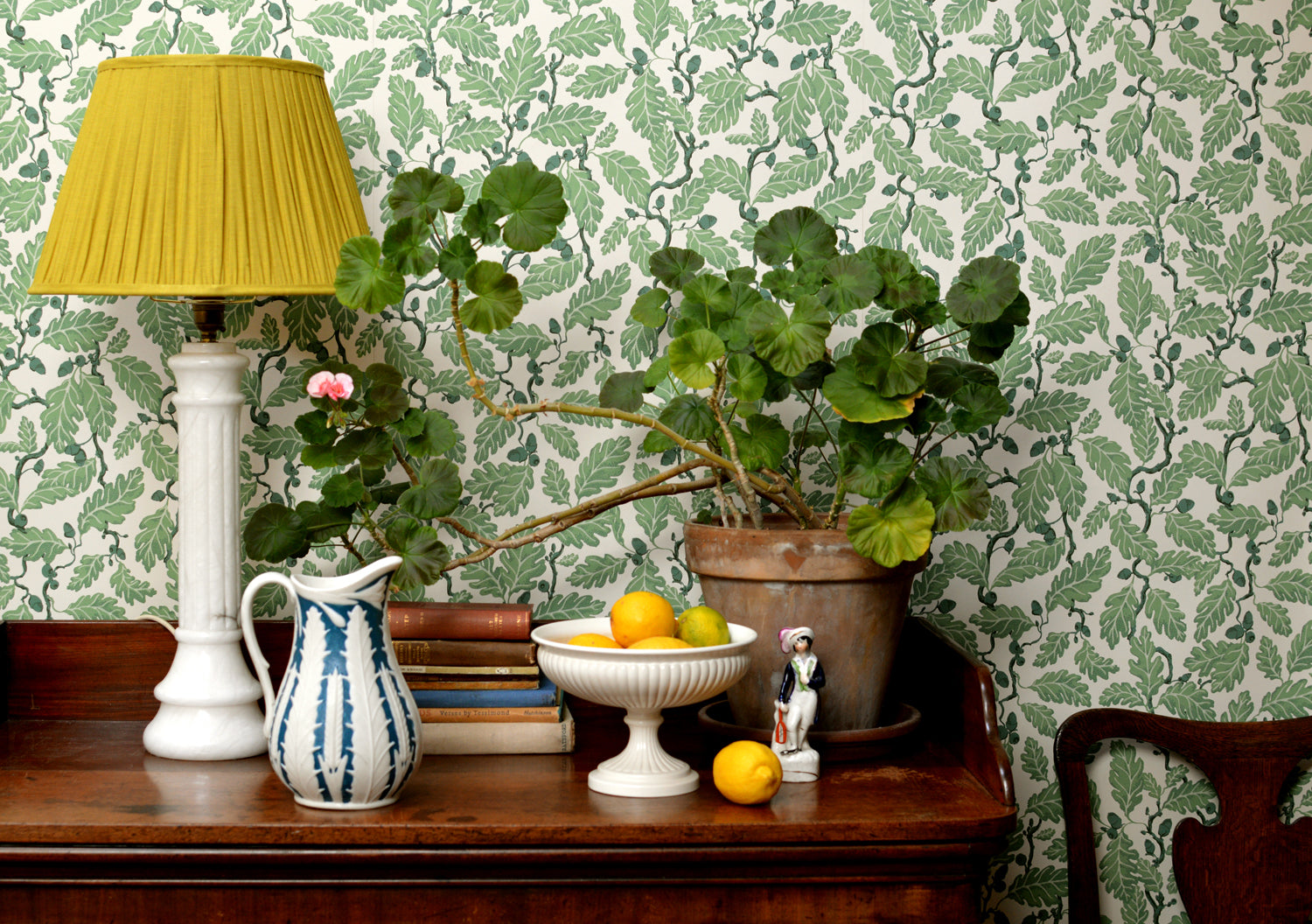 Wooden sideboard with plants, ceramic vesses, lamp and books against a green leaf wallpaper.