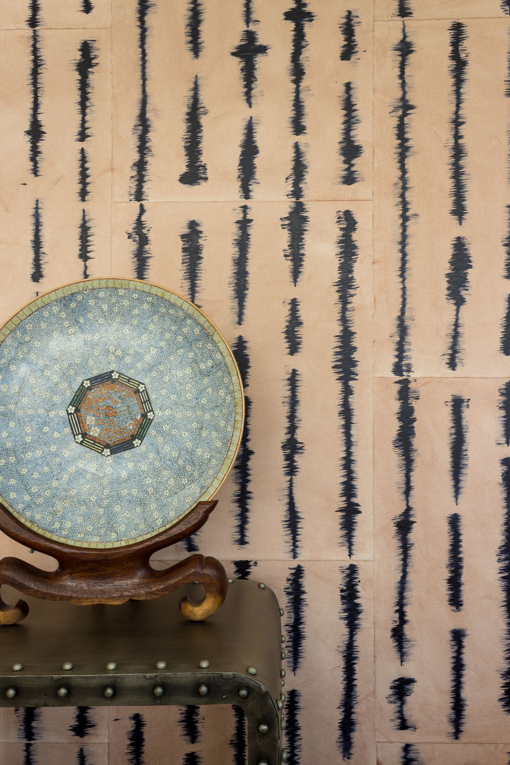 A china plate stands in front of a wall papered in an irregular vibrating linear pattern in navy on a tan field.