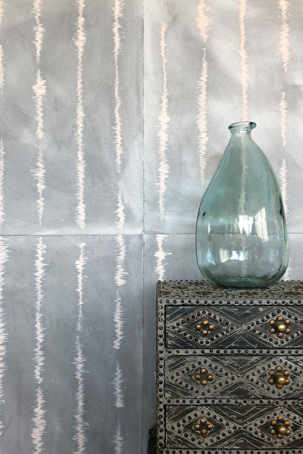A vase on an end table stands in front of a wall papered in an irregular vibrating linear pattern in peach on a greige field.
