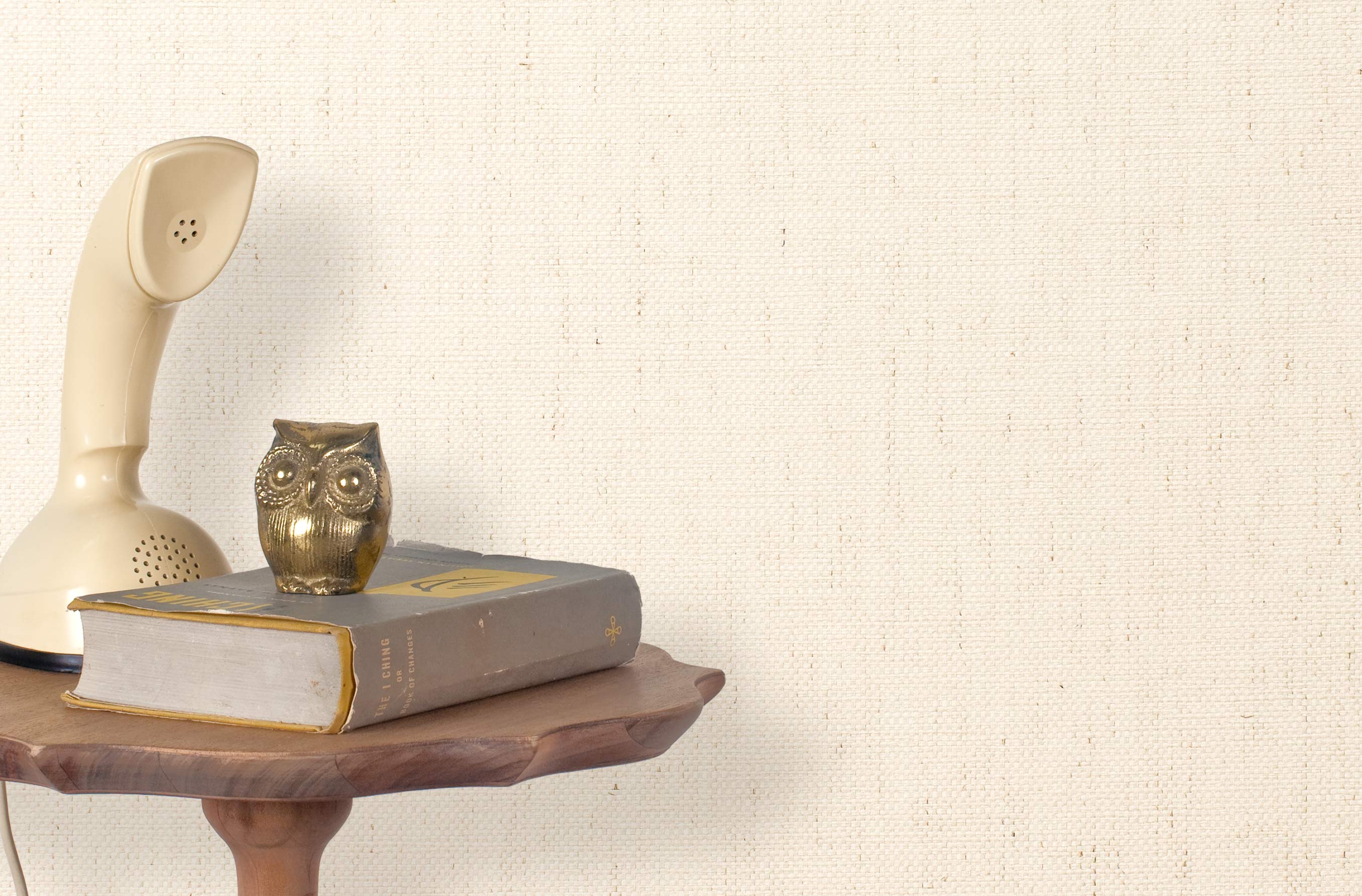 End table with a phone, book and owl sculpture in front of a wall covered in paperweave grasscloth in cream.