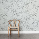 A wooden chair stands in front of a wall papered in a painterly bird and branch pattern in shades of gray on a light gray field.