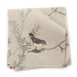 A stack of fabric swatches in a painterly bird and branch pattern in shades of gray on a tan field.