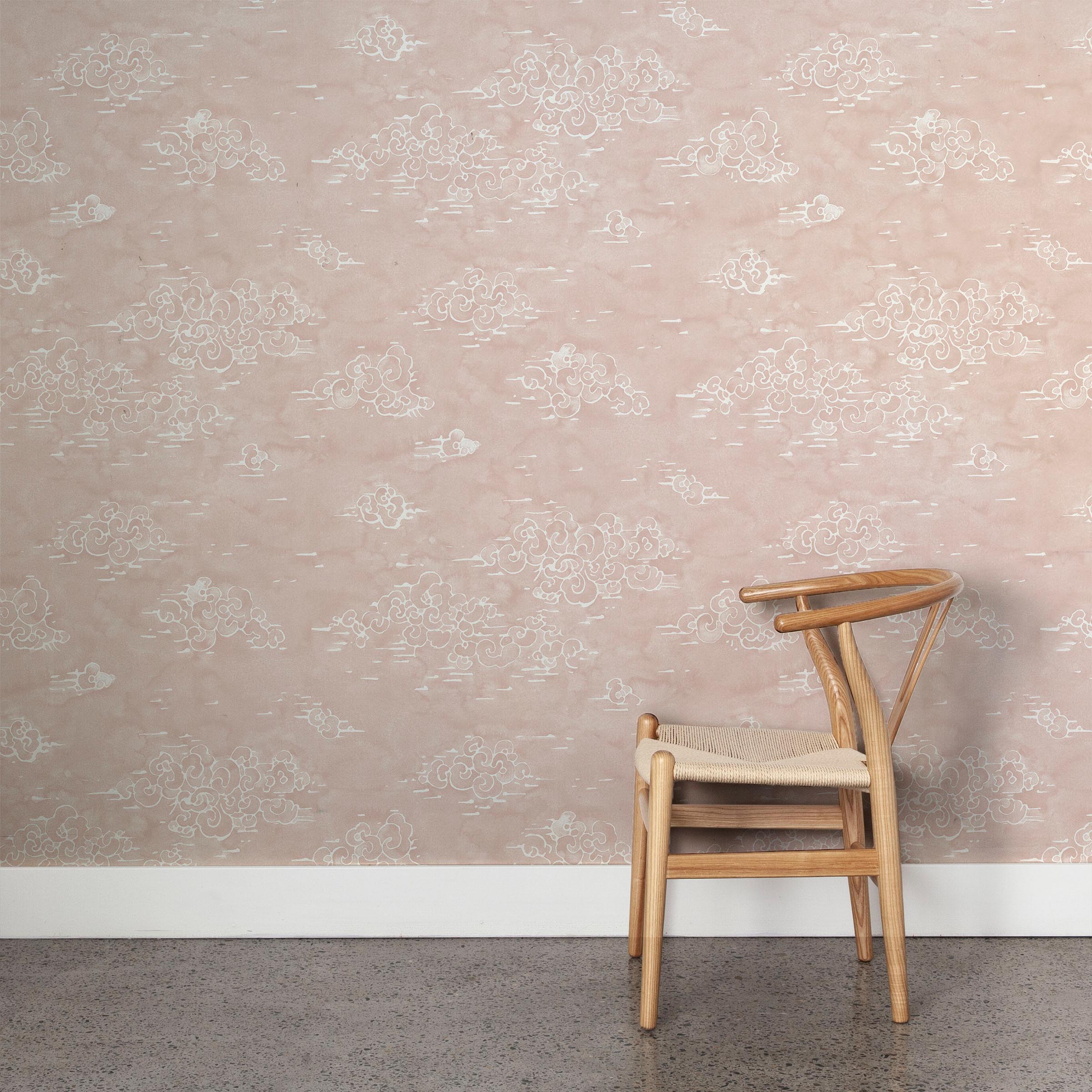 A wooden chair stands in front of a wall papered in a painterly cloud pattern in white on a light pink field.