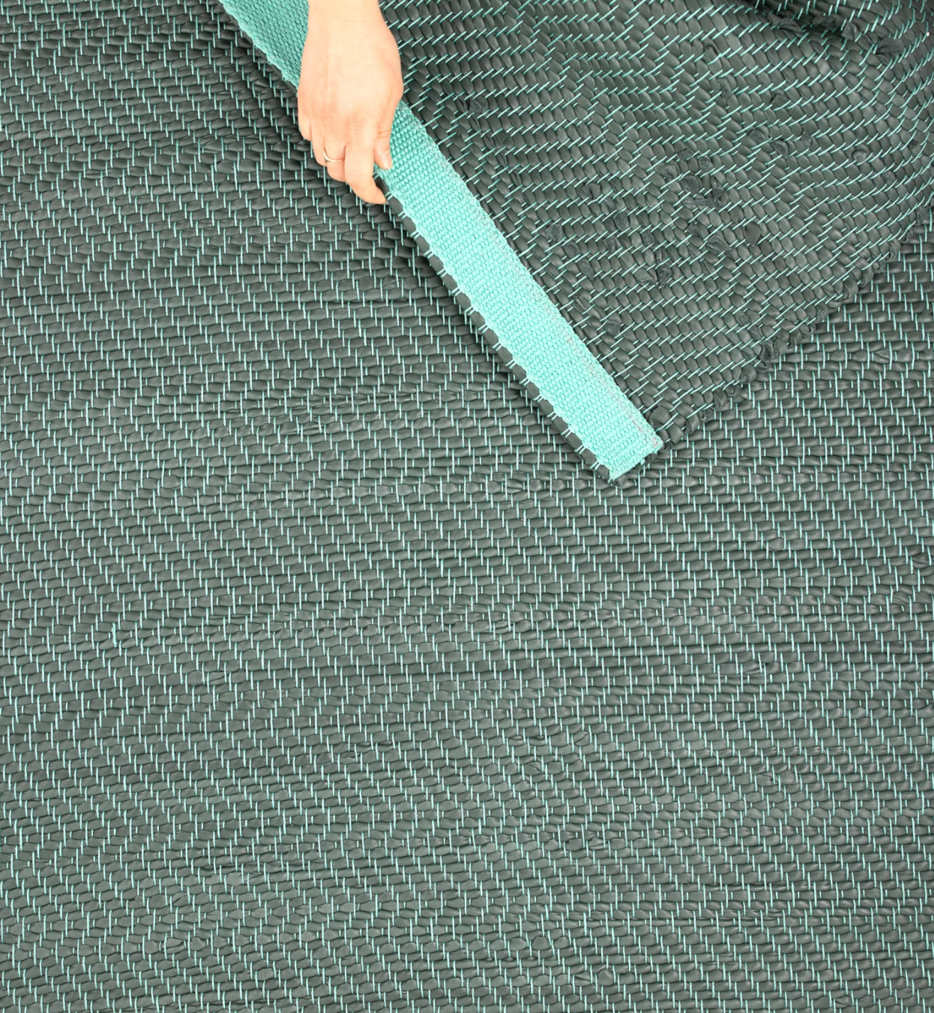 A woman's hand folding a woven leather rug in a herringbone pattern in dark green with turquoise accents.