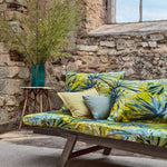 An outdoor space with a sofa upholstered in a painterly palm tree print in shades of yellow, blue and green.