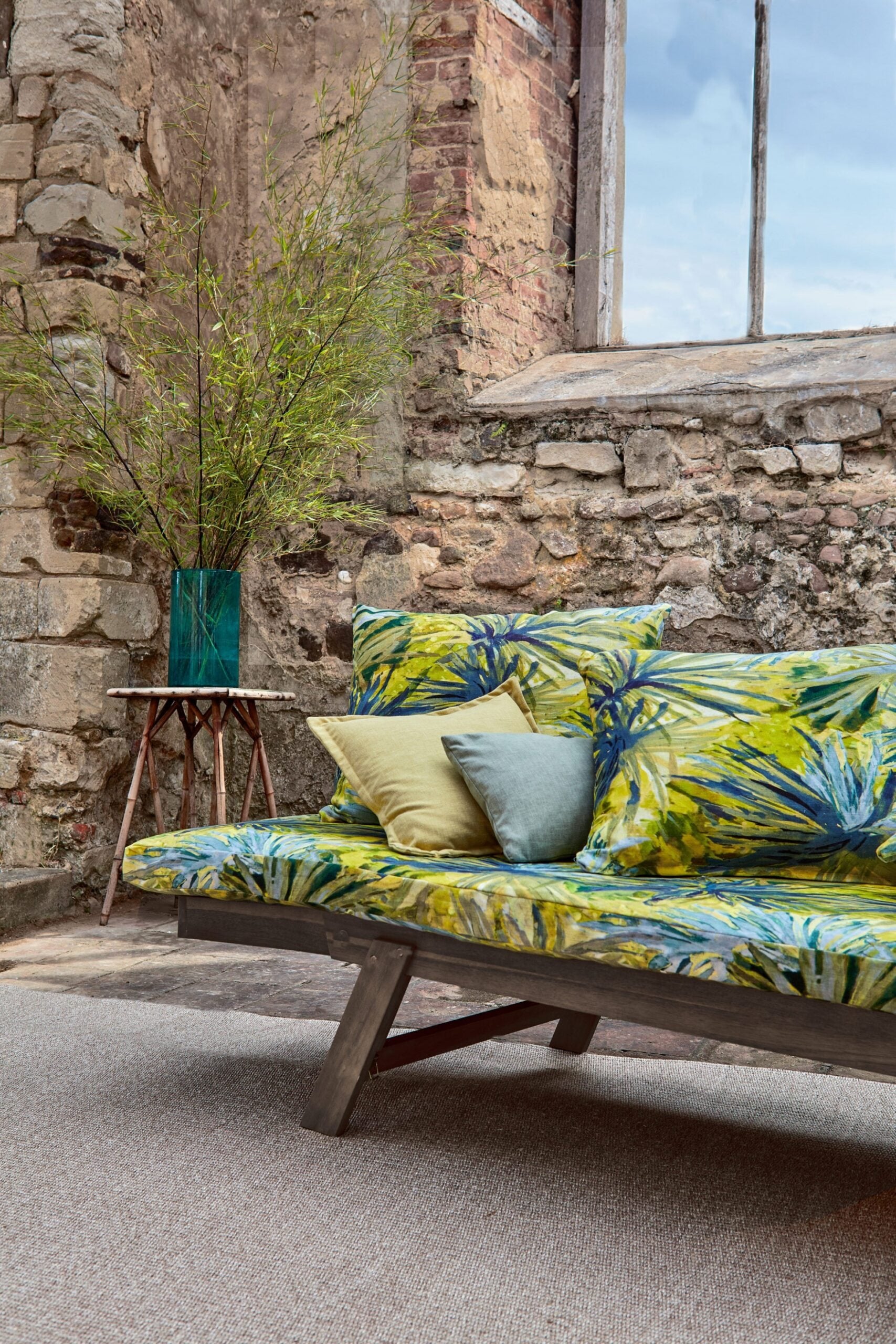 An outdoor space with a sofa upholstered in a painterly palm tree print in shades of yellow, blue and green.