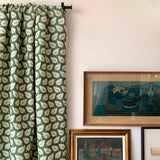 Green and white leaf patterned curtain against a white wall with framed still life paintings in moody colors. 