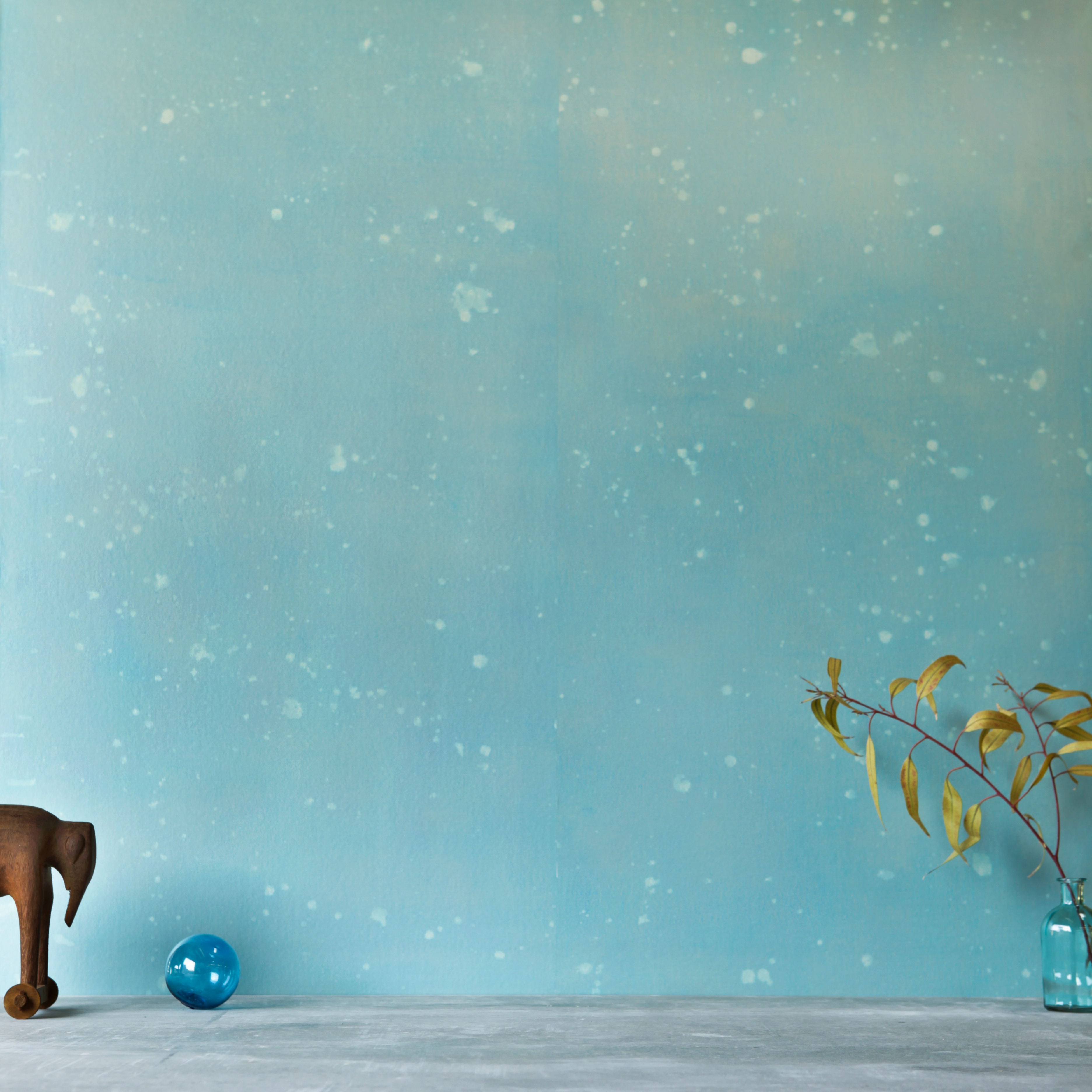 A table with knick knacks stands in front of a wall papered in a blurred paint splatter pattern in blue and turquoise.