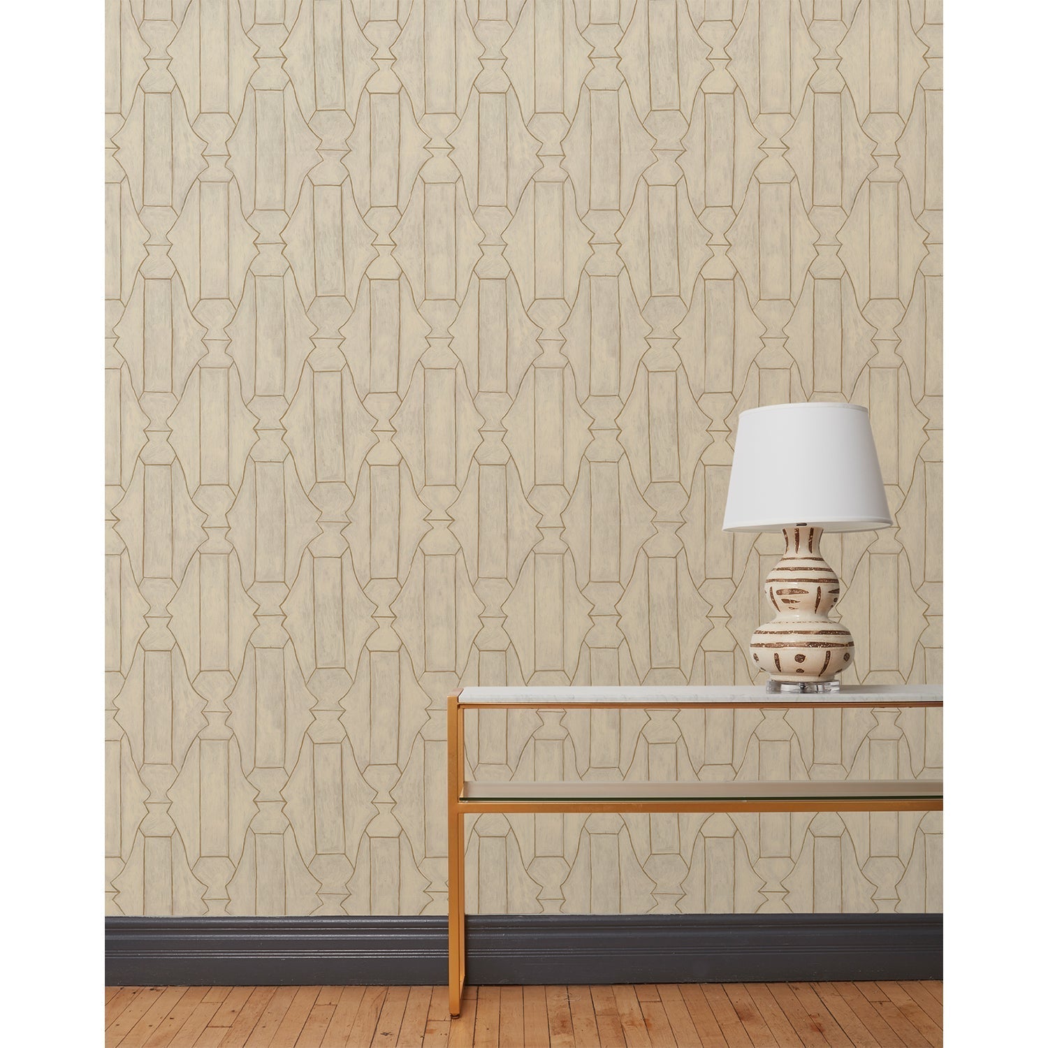 End table and white lamp in front of a wall papered in a repeating curvolinear print in gold on a painted cream field.