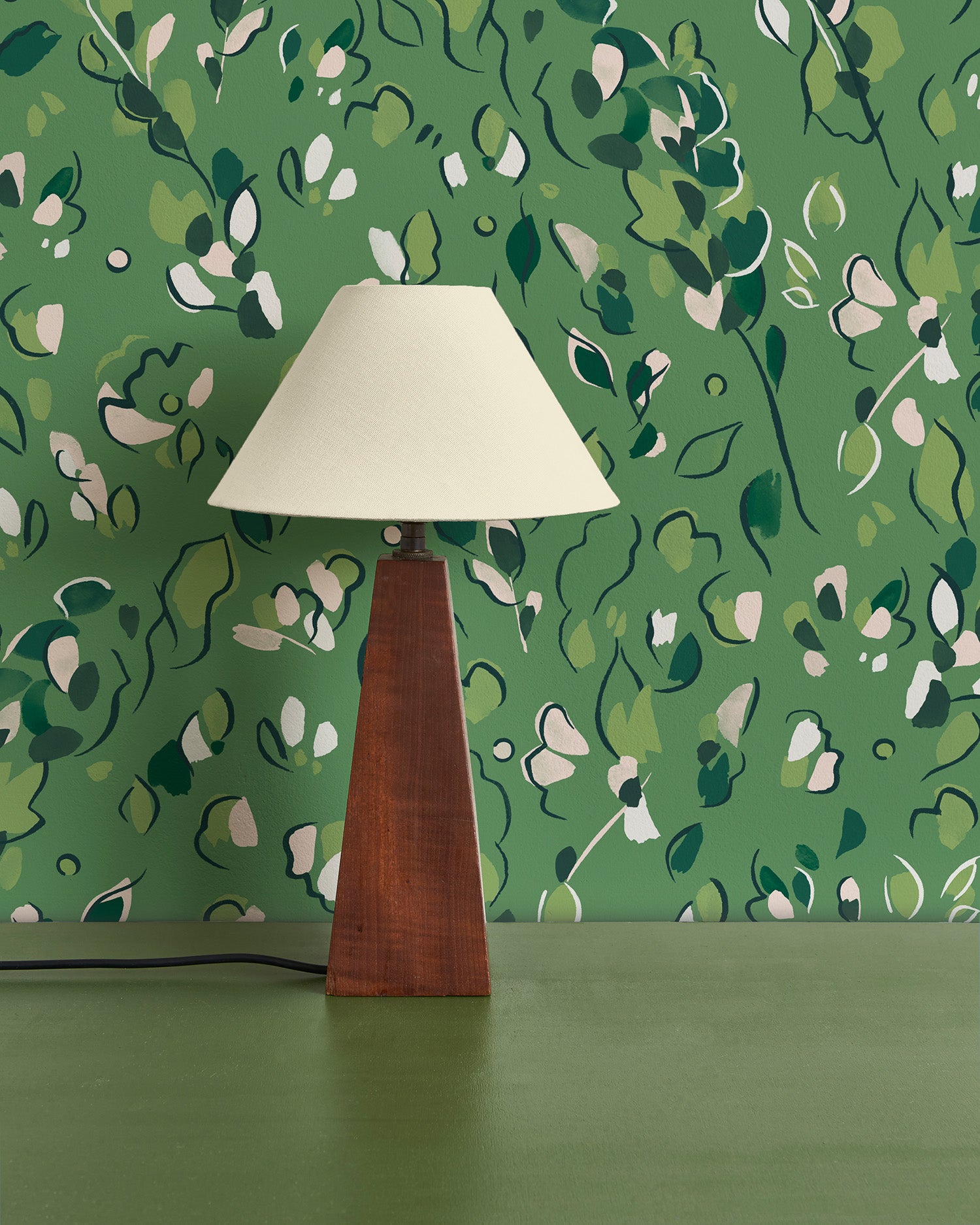 Modernist lamp in front of a wall papered in a painterly leaf print in shades of green.