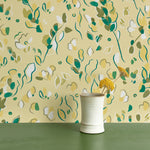 Vase of flowers stands in front of a wall papered in a painterly leaf print in shades of green, white and yellow.