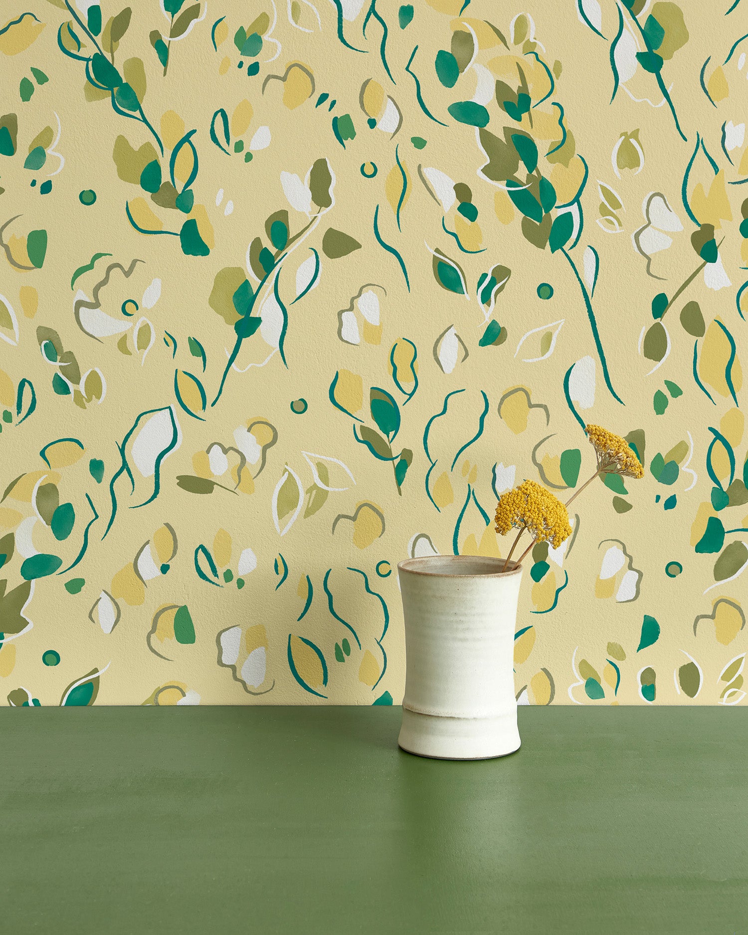 Vase of flowers stands in front of a wall papered in a painterly leaf print in shades of green, white and yellow.