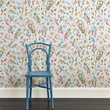 A blue chair stands in front of a wall papered in a painterly leaf print in a rainbow of shades on a white field.