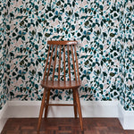 A wooden chair stands in front of a wall papered in a minimal leaf print in shades of pink, turquoise and gray.