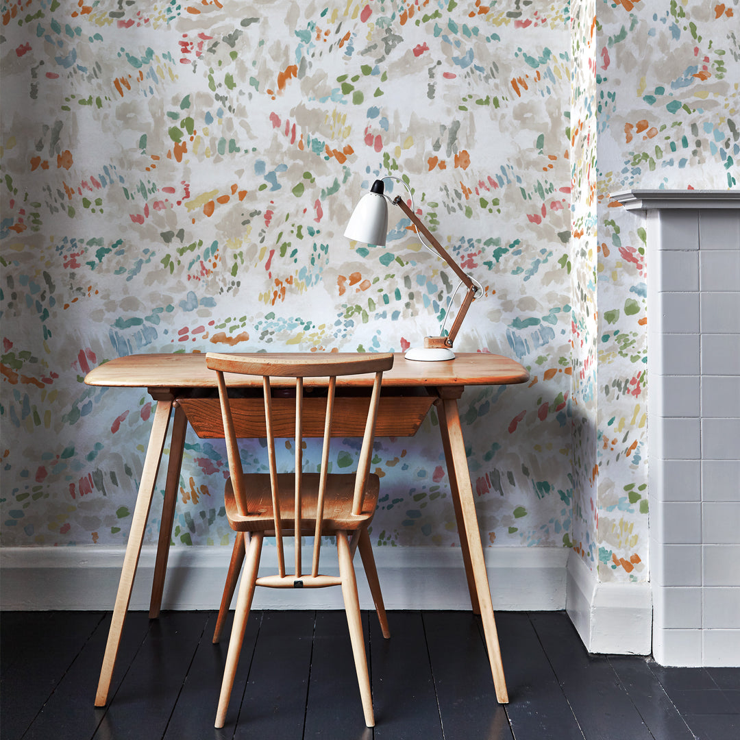 A wooden chair and desk stands in front of a wall papered in a dappled multicolor abstract print.