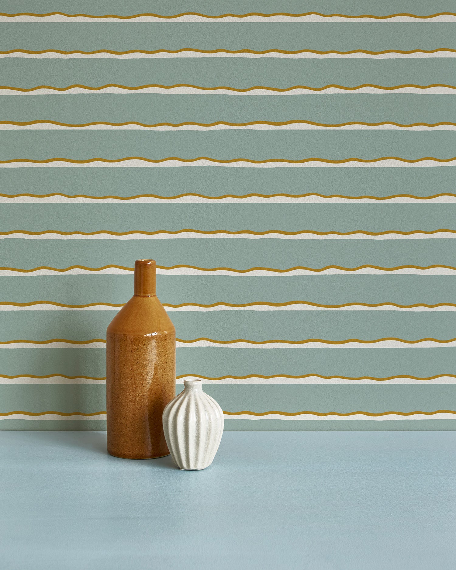 Two vases stand in front of a wall papered in an undulating stripe pattern in gold, white and blue-gray.