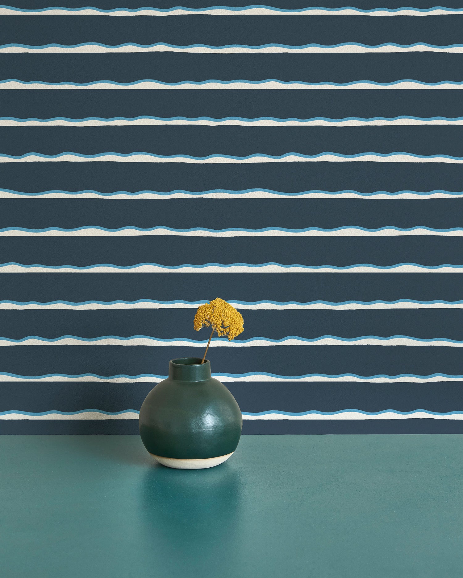 A vase with a flower stands in front of a wall papered in an undulating stripe pattern in blue, white and navy.