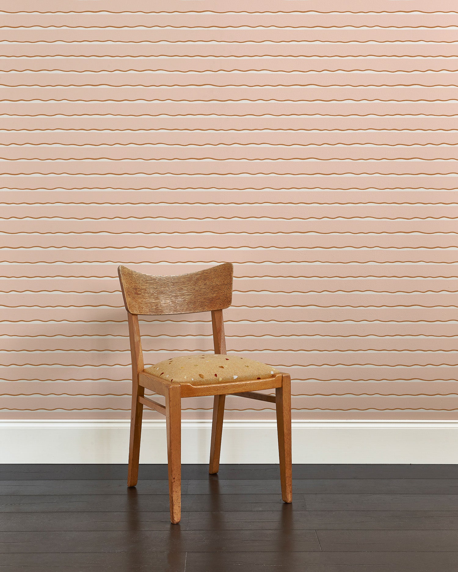A wooden chair stands in front of a wall papered in an undulating stripe pattern in pink, white and rust.