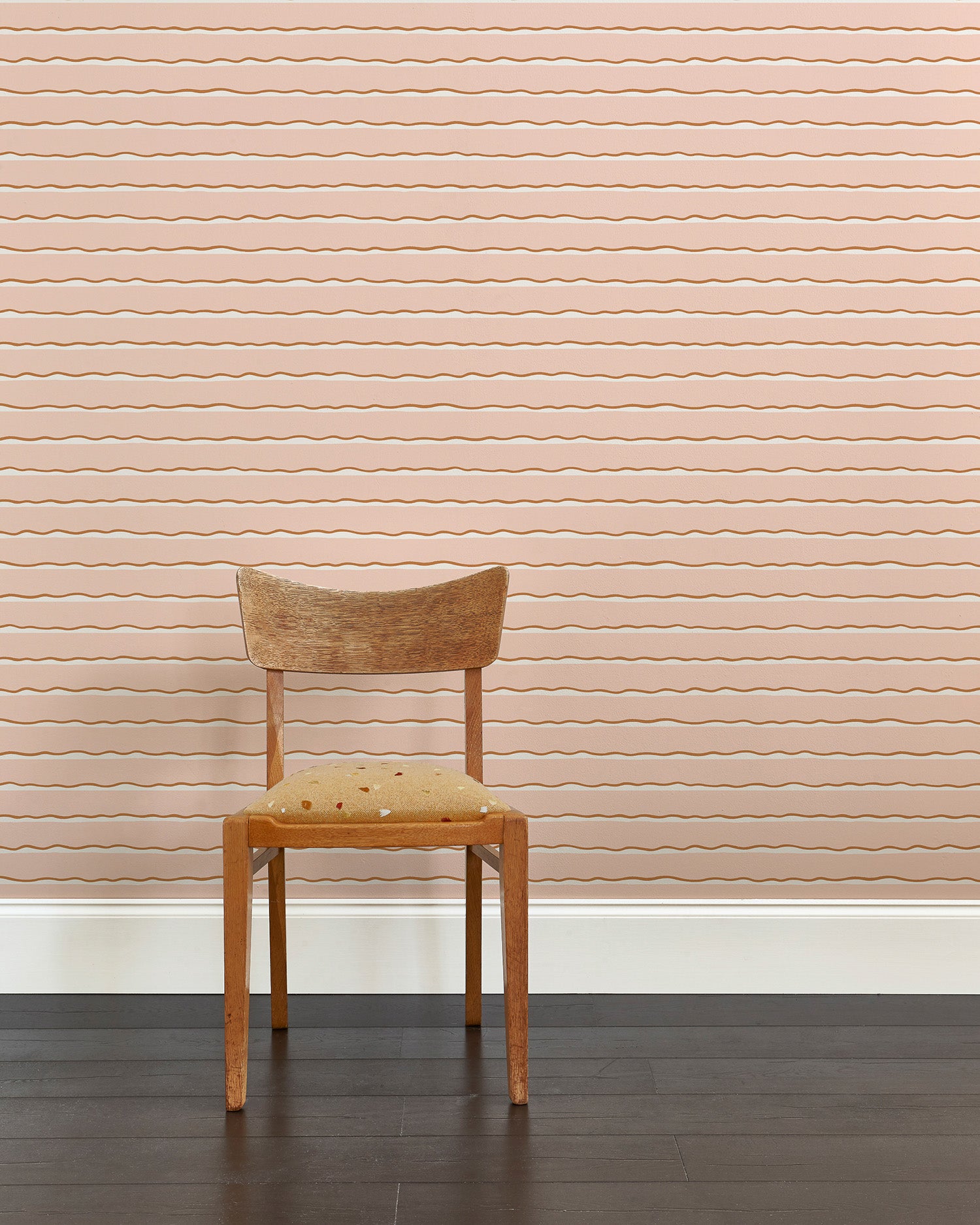 A wooden chair stands in front of a wall papered in an undulating stripe pattern in pink, white and gold.