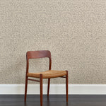 A wooden chair stands in front of a wall papered in a minimalist floral print in gray on a tan field.