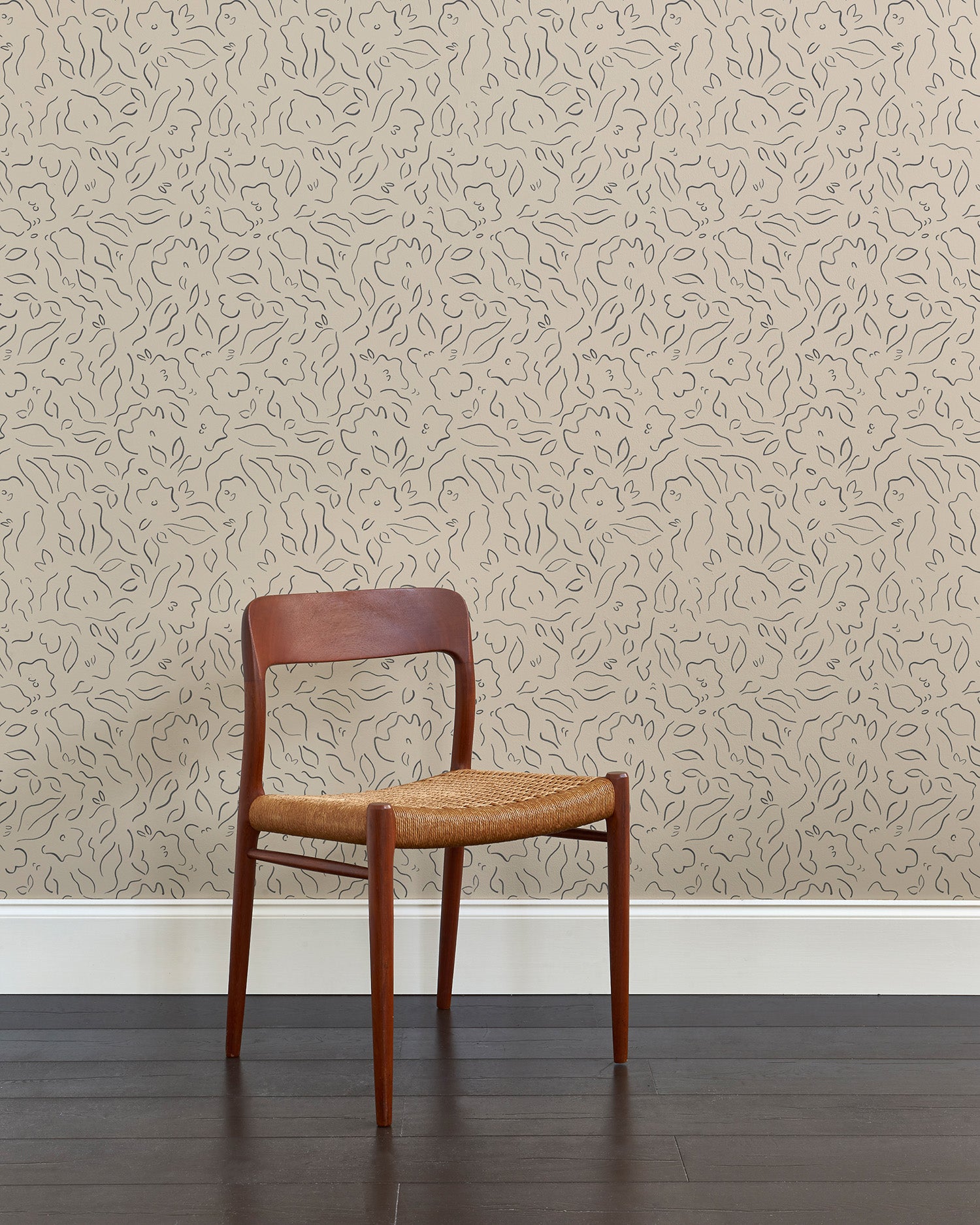 A wooden chair stands in front of a wall papered in a minimalist floral print in gray on a tan field.