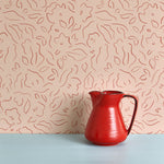 A red vase stands in front of a wall papered in a minimalist floral print in red on a light pink field.