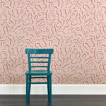 A wooden chair stands in front of a wall papered in a painterly floral print in rust and white on a light pink field.