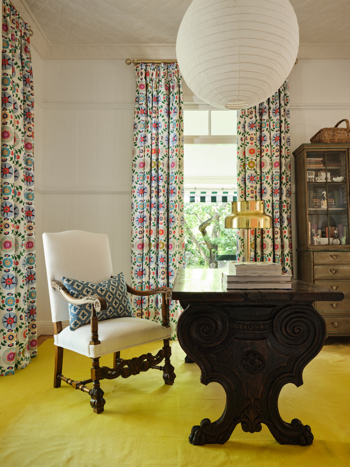 A maximalist study with bright yellow carpet and curtains in a playful rainbow floral print on a white field.