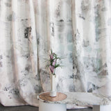 A vase of flowers stands in front of a curtain in a minimal splatter print in shades of gray and pink on a cream field.