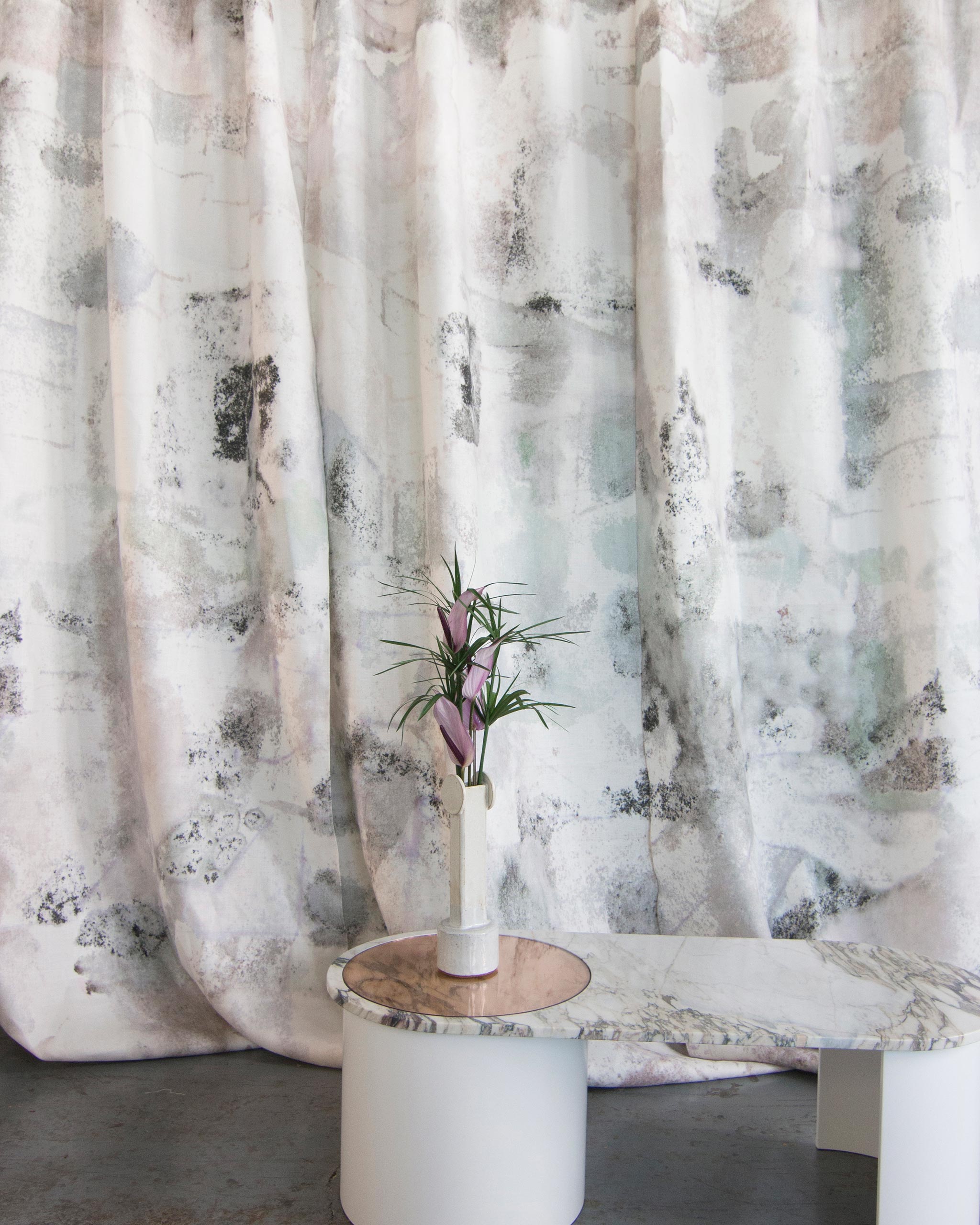 A vase of flowers stands in front of a curtain in a minimal splatter print in shades of gray and pink on a cream field.