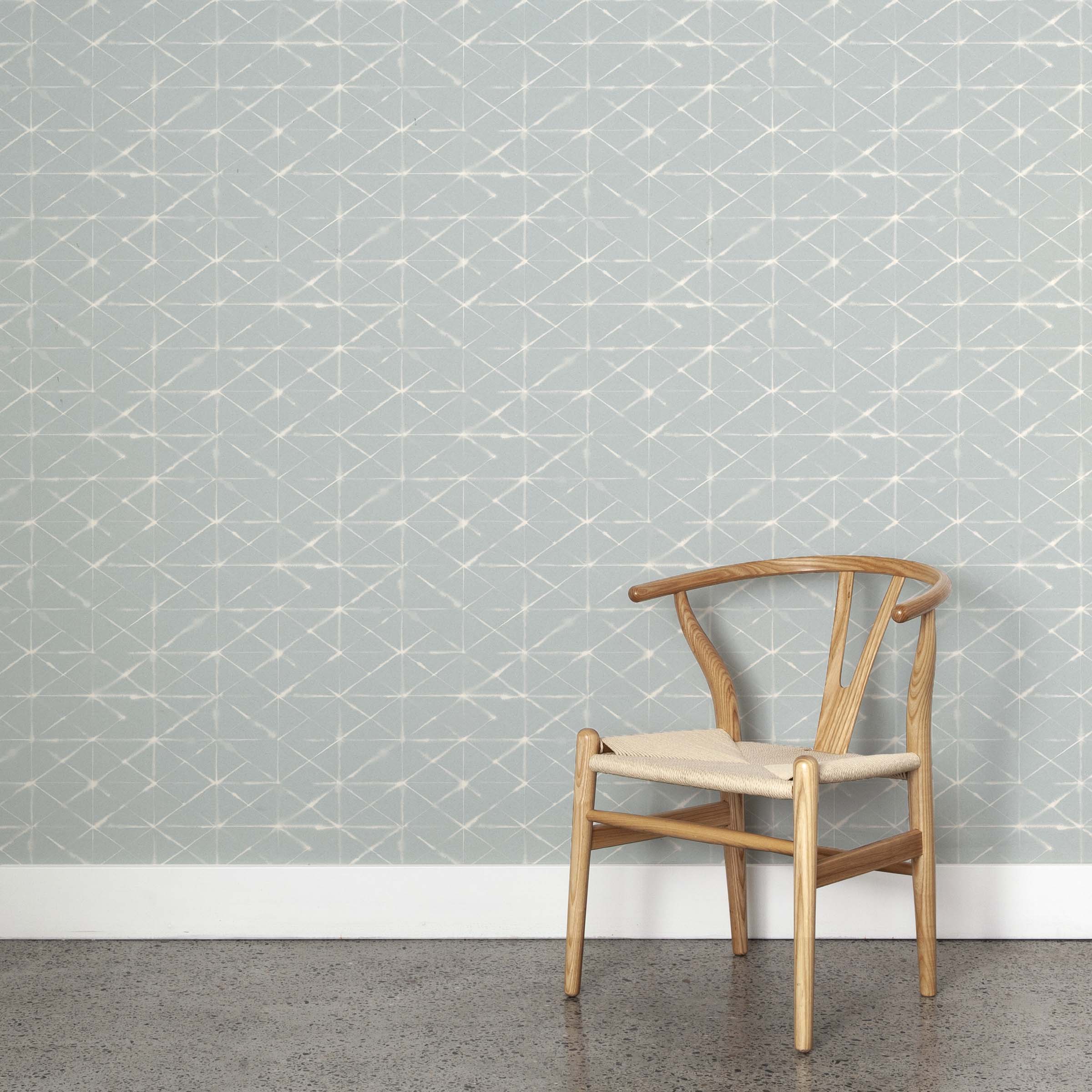 A wooden chair stands in front of a wall papered in a mottled lattice print in white on a light blue field.