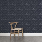 A wooden chair stands in front of a wall papered in a mottled lattice print in white on a black field.