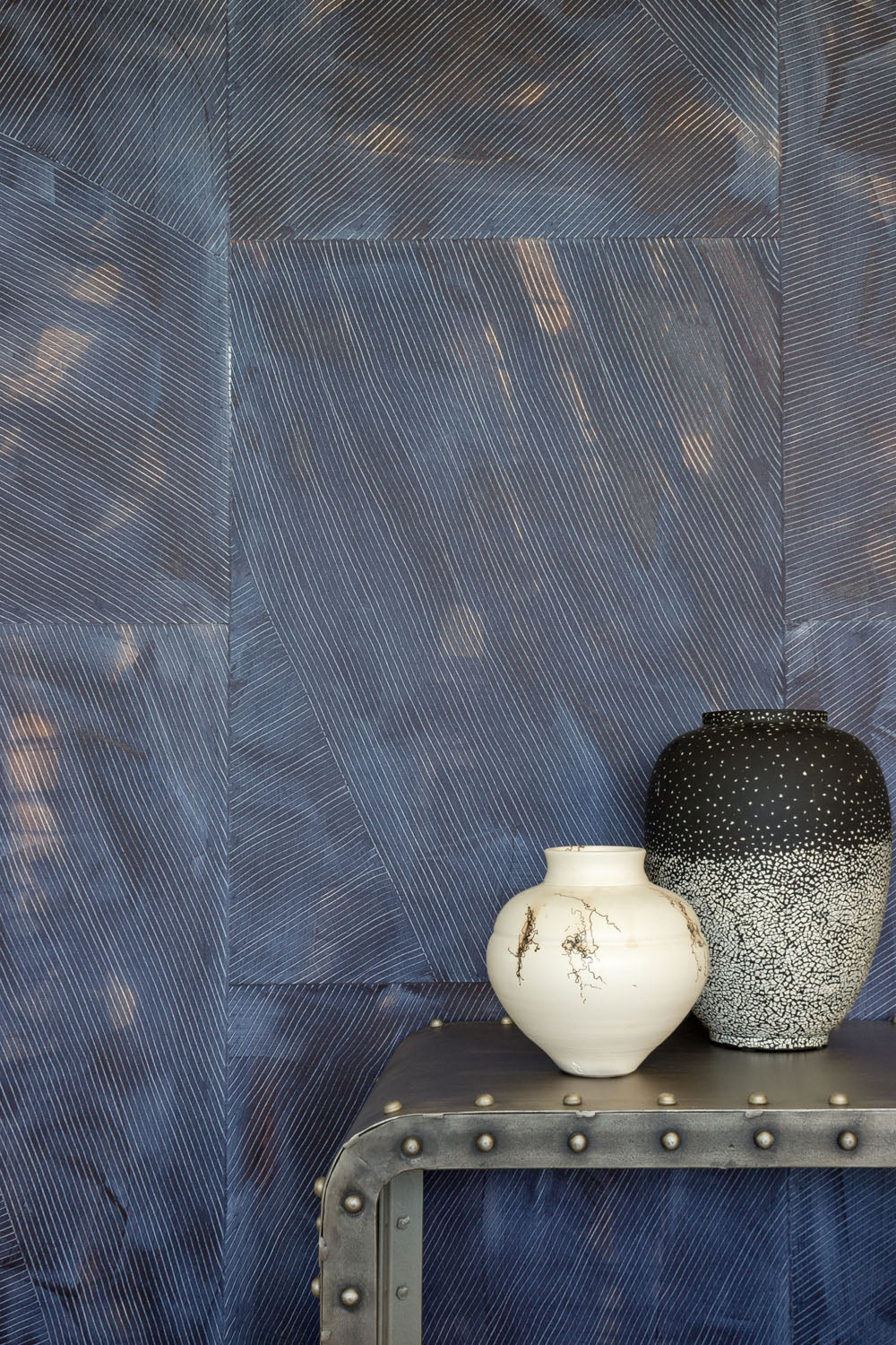 Two vases on a metal end table in front of a wall covered in multi-directional combed wallpaper in mottled indigo.
