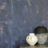 Two vases on a metal end table in front of a wall covered in multi-directional combed wallpaper in mottled indigo.
